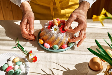 Female hands decorating Easter cake with small colorful chocolate eggs and daisy flowers. Anonymous woman making traditional easter cake or sweet bread with topping. Easter treat.