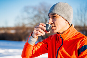 Man drinking tea from mug while standing in nature in winter, man in orange jacket and hat enjoying cup of coffee in winter forest,