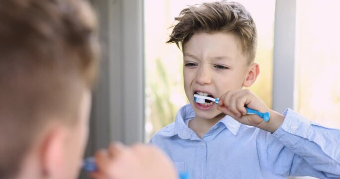 Close up cute little boy looking in mirror doing morning routine brushing teeth with toothpaste and soft manual toothbrush in bathroom at home. Caring for dental health and wellness, oral self-care