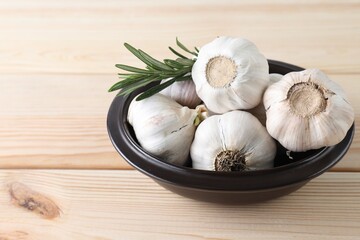 Fresh garlic bulbs in bowl and rosemary on wooden table, closeup. Space for text