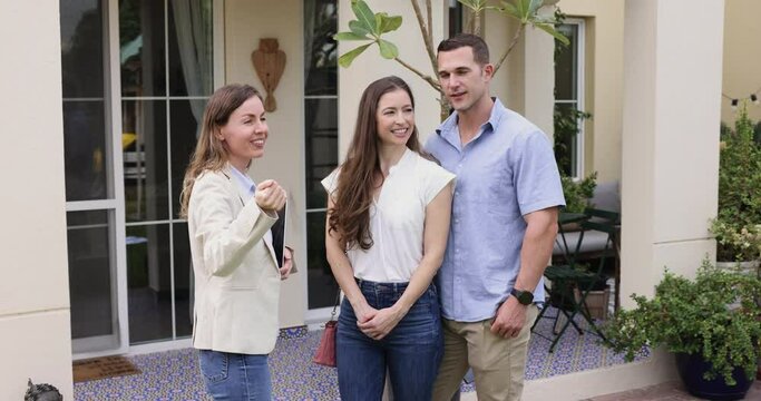 Female real-estate agent showing to young couple property, convincing to buy house, talking about benefits and facilities standing threesome in country house backyard. Housing improvement, bank loan