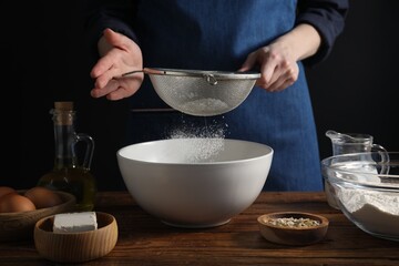 Making bread. Woman sifting flour over bowl at wooden table on dark background, closeup