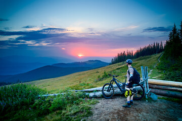 Cyclist man riding electric bike outdoors in the evening. Male tourist resting on grassy hill,...