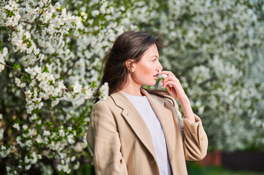 Woman allergic using medical nasal spray, suffering from seasonal allergy at spring in blossoming garden. Young woman treating runny nose in front of blooming tree outdoors. Spring allergy concept. - Powered by Adobe