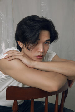 A young Asian man with dark hair in casual white T-shirt sits on wooden chair, resting arms across chairs back. He gazes intently toward the camera, within a softly lit studio.