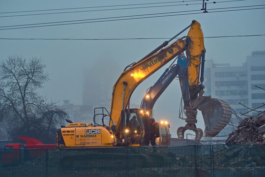 Minsk, Belarus. Feb 11, 2024. JCB and Hitachi excavators with scoop and grapple removes waste after building demolishion on construction yard. Removing rubble from demolition yard early in the morning