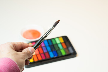 A hand holds a wet brush on a warm colored watercolor kit.