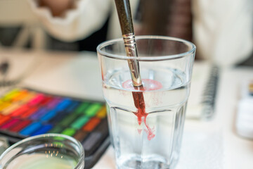 Detail of a watercolor brush dropping red watercolor pigment into a glass of water.