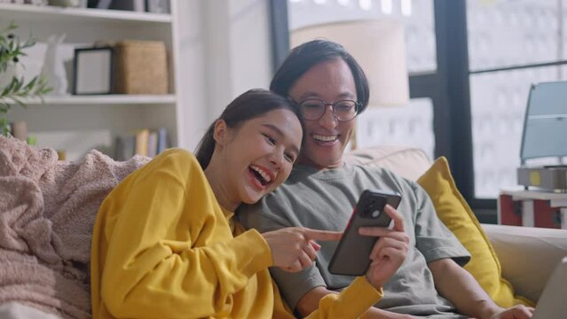 Asian couple family lifestyle,technology concept.Happy young Asian couple using cellphone,watch movie online,browsing internet,chatting online,checking new mobile application,relaxing on sofa at home