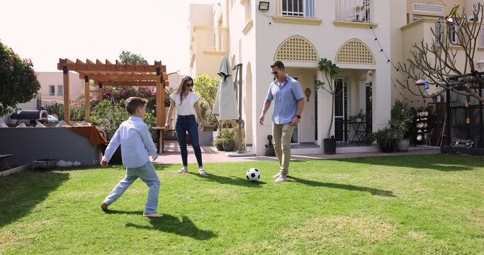Married couple with little 8s son play football in backyard of their modern country house, spend sunny summer warm day engaged together in playtime, hobby and active leisure outdoors. Family pastime