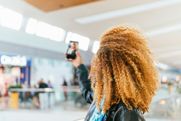 An unrecognizable young black woman with curly hair is taking a picture of herself with a cell phone. She is wearing a black jacket and a blue scarf. The scene takes place in a shopping mall