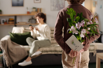 Rear view of little boy with bunch of fresh flowers and handmade postcard for mother day standing...