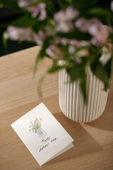 Part of wooden table with handmade postcard with greetings for mother day and drawing of bunch of red tulips in vase