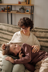 Happy young brunette woman in eyeglasses enjoying play with her adorable son while both relaxing on couch on weekend