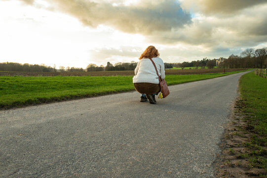 Auburn haired woman seen crouching in the middle of an empty road with the aim of taking landscape picture in early spring in the UK.