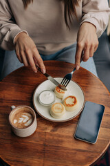 An unrecognizable woman using cutlery has breakfast in a cafe with cheese pancakes and drinks coffee.
