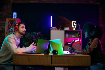 Young people, social media influencers sitting at table with microphones and laptops, recording...