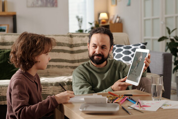 Bearded father with tablet sitting by small table and showing his son example of postcard for...