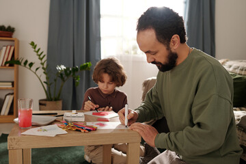 Bearded man with highlighter bending over small table and drawing something on paper against his...