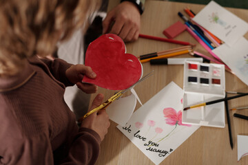 High angle of hands of child cutting out red paper heart over table with crayons, handmade...