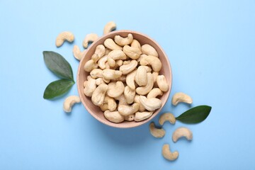 Many tasty cashew nuts and leaves on light blue background, top view