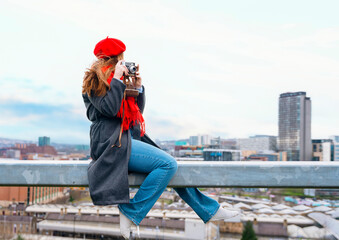 happy  woman in a ghrey coat, red scarf and beret  sitting in front of the city Sheffield  and take photos on great spring day