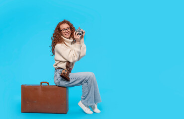 Amazing  redhead woman in a white sweater sitting on a suitcase, holding old camera and dreaming about a journey on a blue background with copy space.  Travel concept