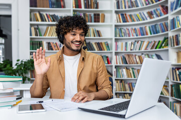 A young man with curly hair and a headset smiles and waves at his laptop camera, engaged in an online video call while seated at a desk in a modern library setting with books in the background.