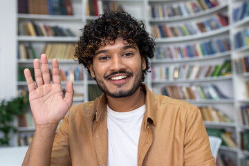 A smiling man with curly hair waves to the camera, exuding warmth and friendliness, in a relaxed...