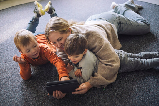 High Angle View Of Kids Watching Digital Tablet With Female Teacher Lying Down On Carpet At Preschool