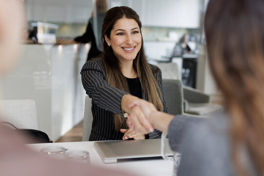 Happy saleswoman greeting female customer with handshake while sitting at desk in real estate office