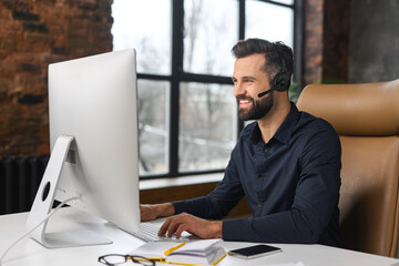 A charismatic businessman wearing headphones and a smart casual shirt is sitting at a desk in front of a personal computer, involved virtual meeting - obrazy, fototapety, plakaty
