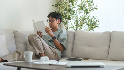 Calm asian man in casual clothes sitting on couch in bright living room and reading book.