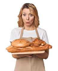 Middle-aged Caucasian woman baker with bread tray shrugs shoulders and open eyes confused.