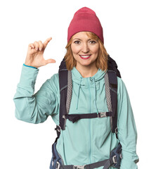 Middle-aged Caucasian woman with hiking gear holding something little with forefingers, smiling and confident.