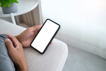 Man using mobile phone while lying on couch at home. White empty screen for advertising.