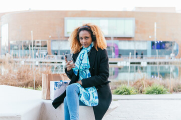 A woman with a headphones is sitting on a bench in shopping mall outdoors. She is holding a cell phone in her hand and listening music while rest of a shopping day