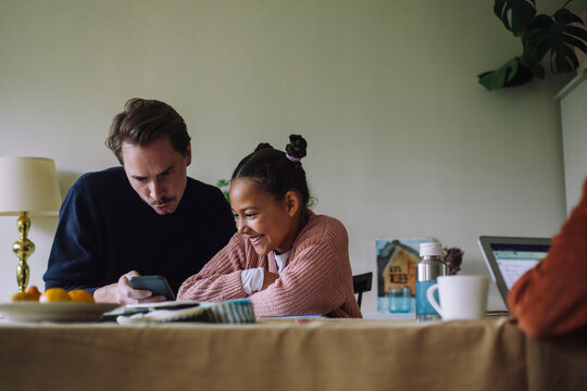 Gay man sharing smart phone with happy daughter sitting at dining table in home