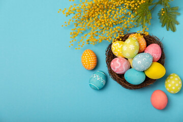 Easter eggs in the nest with mimosa flowers on a bright yellow background. Easter celebration concept. Colorful easter handmade decorated Easter eggs. Place for text. Copy space.