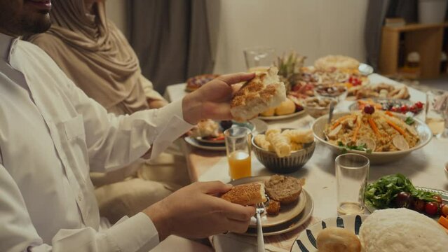 Side tilt footage of smiling bearded muslim man sharing flatbread or pita with family members during festive dinner celebrating Eid al-Fitr at home