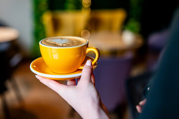 A person holds a vibrant yellow coffee cup with latte art in a cozy café setting. Warm lighting and potted plants create an inviting atmosphere