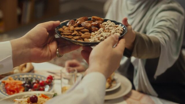 Close up of anonymous muslim woman in hijab passing plate with almond, dates and chickpeas over table during festive dinner with family on occasion of celebrating Eid al-Fitr