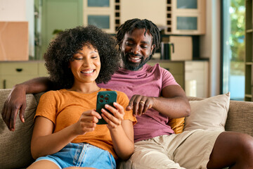 Couple Sitting On Sofa At Home Looking At Mobile Phone Together