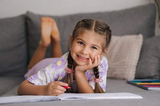 Adorable little girl with beautiful braid hairstyle lying on sofa at home and drawing picture for mom. Girl look into camera