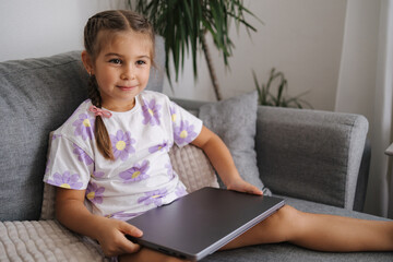 Adorable little girl take father's laptop and sits on sofa at home. 