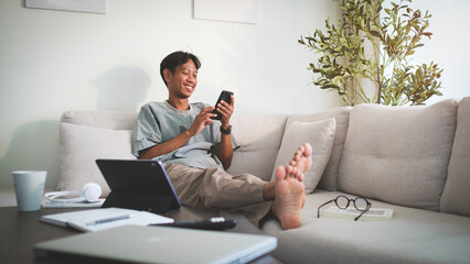 Handsome asian man reading messages on mobile phone while sitting on couch in cozy living room. - obrazy, fototapety, plakaty