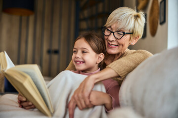 Smiling grandma and her grandchild reading a book at home while sitting on the couch, covered in a...