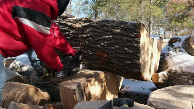 Slow motion of man in red jacket in hood using gas powered chainsaw to cut oak tree logs into smaller pieces to be processed into firewood.