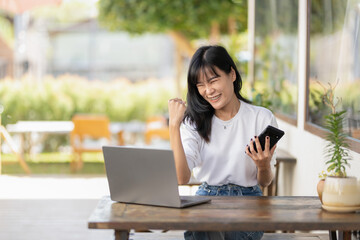Happy Asian woman raising fist in victory while looking at her smartphone with a laptop in an outdoor cafe