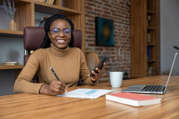 Satisfied woman with dreadlocks working with laptop and smartphone coordinates corporate tasks